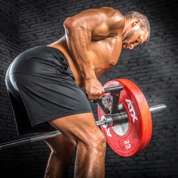 A muscular person in black shorts is performing a bent-over row with an ATX Parallel Grip T Bar Row Handle and a 7ft 1500lbs Olympic Bushing Bar, loaded with red weight plates, against a gyms gray brick wall backdrop.