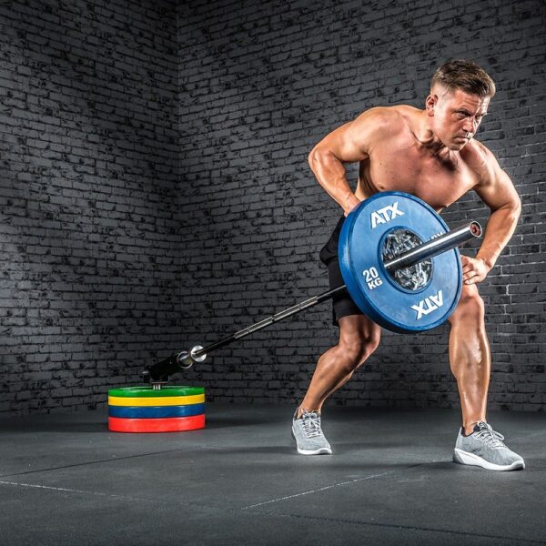 A shirtless muscular man works out in a gym, using an ATX® Single Arm Row Barbell Handle and a 20 kg weight plate. He wears black shorts and gray sneakers. Stacked colorful weight plates are behind him against a brick wall.