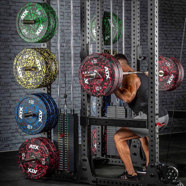 A person in a gym squats with a barbell loaded with ATX® weight plates. The background displays a rack of colorful weight plates.