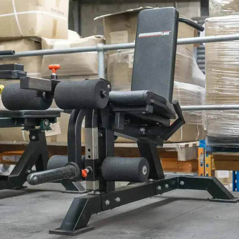 The ATX Leg Extension Leg Curl Machine in the gym has a black padded seat and backrest with foam rollers for leg support. Stacked cardboard boxes and pallets in the background add an industrial feel.