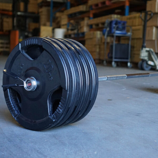 The Power Maxx 6ft Olympic Barbell, stacked with multiple 25 kg plates on both sides, sits on the gym floor. The backdrop features shelves and assorted equipment, highlighting a focused weightlifting or fitness area.