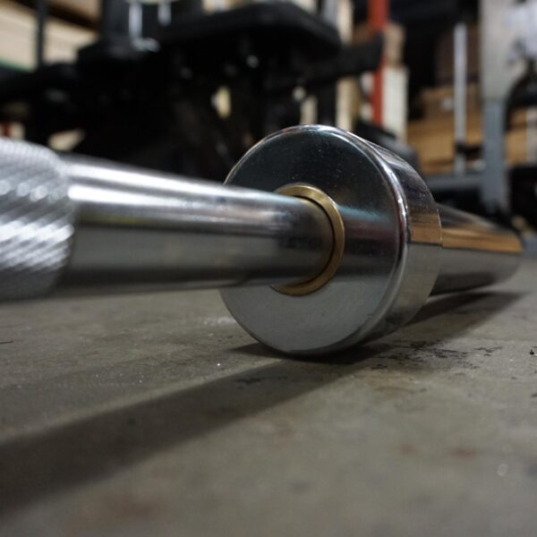 A close-up shows the 7ft Olympic Barbell Thrust Bearings 1500lbs resting on a gym floor. The shiny metal finish of the barbell end reflects the light, while blurred gym equipment and weights fill the background.