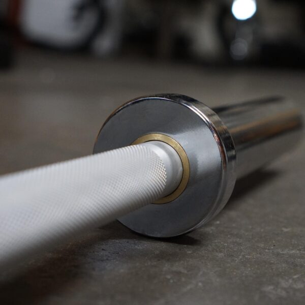 Close-up of the Aluminium 7.5kg Technique Olympic Bar resting on a gym floor. The textured grip of the metal bar contrasts with its gleaming, smooth ends. The focus is on the end part of the bar, with the background slightly blurred.