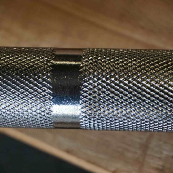 Close-up of a 7ft Olympic Barbell Thrust Bearings 1000lbs with a smooth, shiny band around the knurled middle for grip. The background is blurred, featuring a wooden surface.