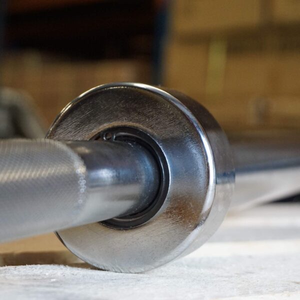 Close-up of the 7ft Olympic Barbell Thrust Bearings 1000lbs, highlighting the knurled grip and metallic weights. Detailed texture of the barbell is in focus within a gym setting, with a blurred background hinting at other equipment.