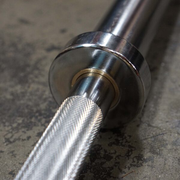 Close-up of the Power Maxx Big Olympic Dumbbell Handle on a textured concrete floor, showcasing its metallic sleeve and intricate knurled grip. The shiny finish reflects light, highlighting superior grip detailing.