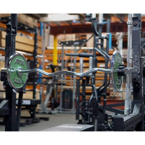 In a gym filled with weightlifting gear, a barbell on the rack uses green ATX weight plates alongside an Olympic EZ Curl Bar, with shelves and workout machines visible in the background.