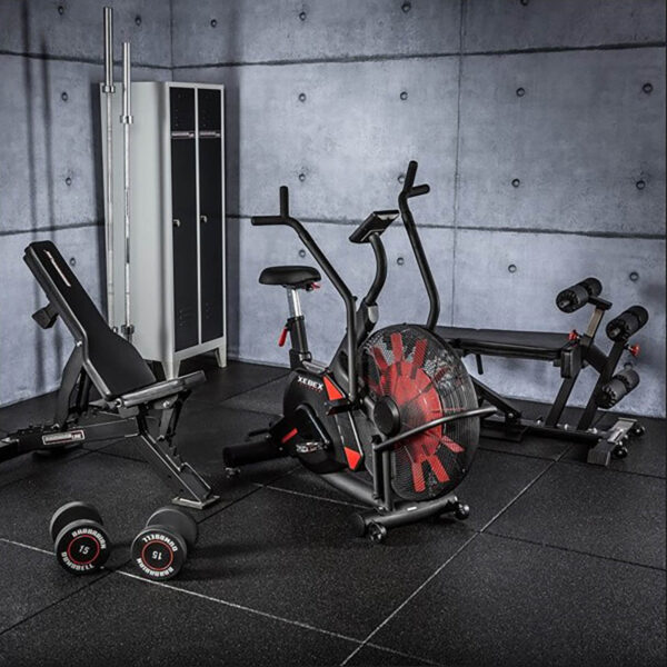 A modern gym room equipped with a stationary bike, weight bench, treadmill, and dumbbells. The concrete walls feature a locker in the background. The floor is covered with GymFloor® 15mm 1m x 1m Black rubber mats.