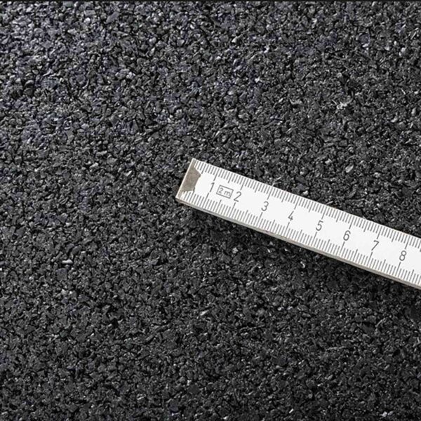 A close-up image of the GymFloor® 15mm 1m x 1m Black surface with a metallic ruler placed diagonally. The ruler, marked in centimeters and partially visible, accentuates the texture and granularity similar to asphalt.