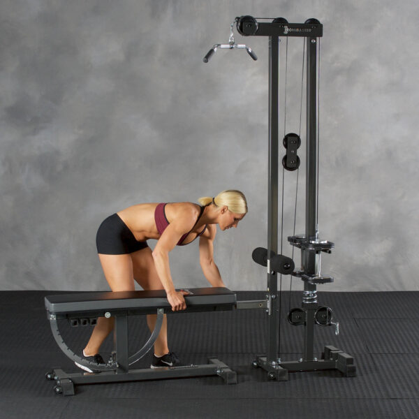 A person in athletic wear adjusts an Ironmaster Cable Tower Attachment V2 next to a workout bench in a gym with gray walls and black mats on the floor.