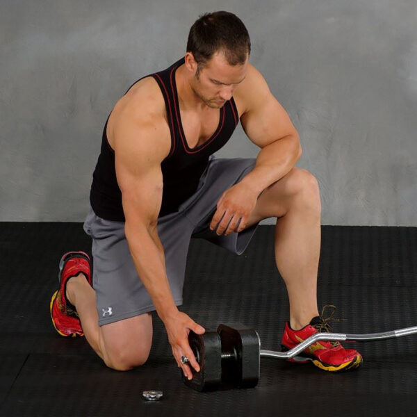 A man in a black tank top and gray shorts kneels on a black mat, adjusting the weight on a dumbbell. Hes wearing red and yellow athletic shoes next to an Ironmaster EZ Curl Bar with a metal weight disk nearby. The background is a plain gray wall.
