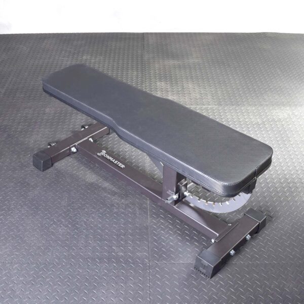 The Ironmaster Hybrid Bench Pad, a black adjustable workout bench with a cushioned pad, rests on a textured gym floor. It is set in a flat position, showcasing visible branding on its sturdy frame.