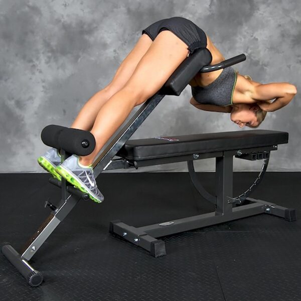Wearing athletic gear, someone uses the Ironmaster Hyper Core Attachment for a back extension exercise. Bent forward with hands behind their head, they are supported by the equipment. The setting includes a gray wall and black floor.