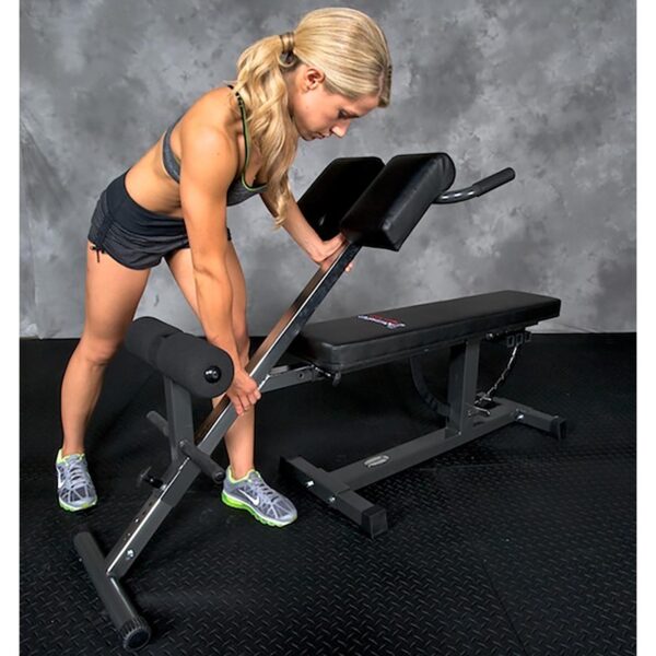 A person with long blonde hair exercises on an Ironmaster Hyper Core Attachment in a gym, wearing a black sports bra, black shorts, and gray sneakers. The background has a gray textured wall and black rubber flooring.