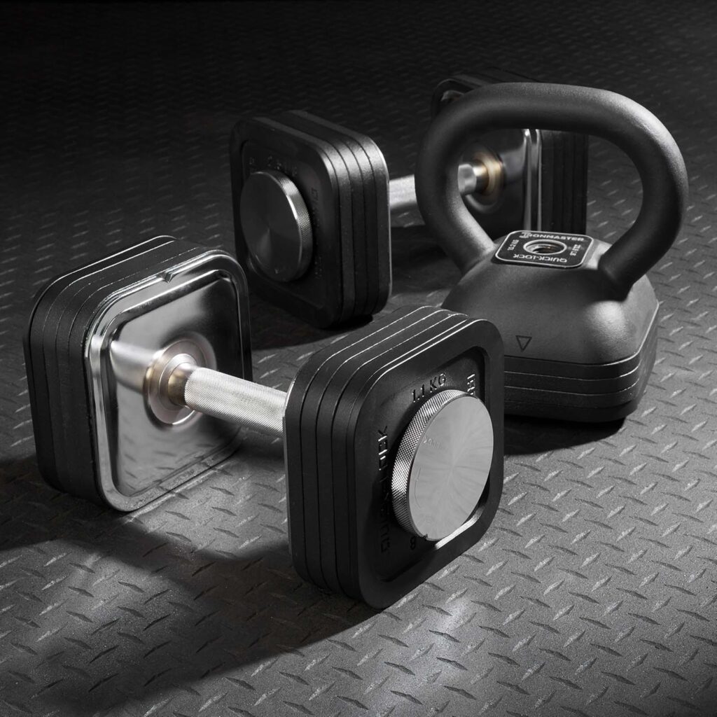 A set of adjustable dumbbells with black square plates and the Ironmaster Adjustable Kettlebell Handle rests on a textured rubber gym floor. The equipment features a modern design, ideal for strength training exercises.