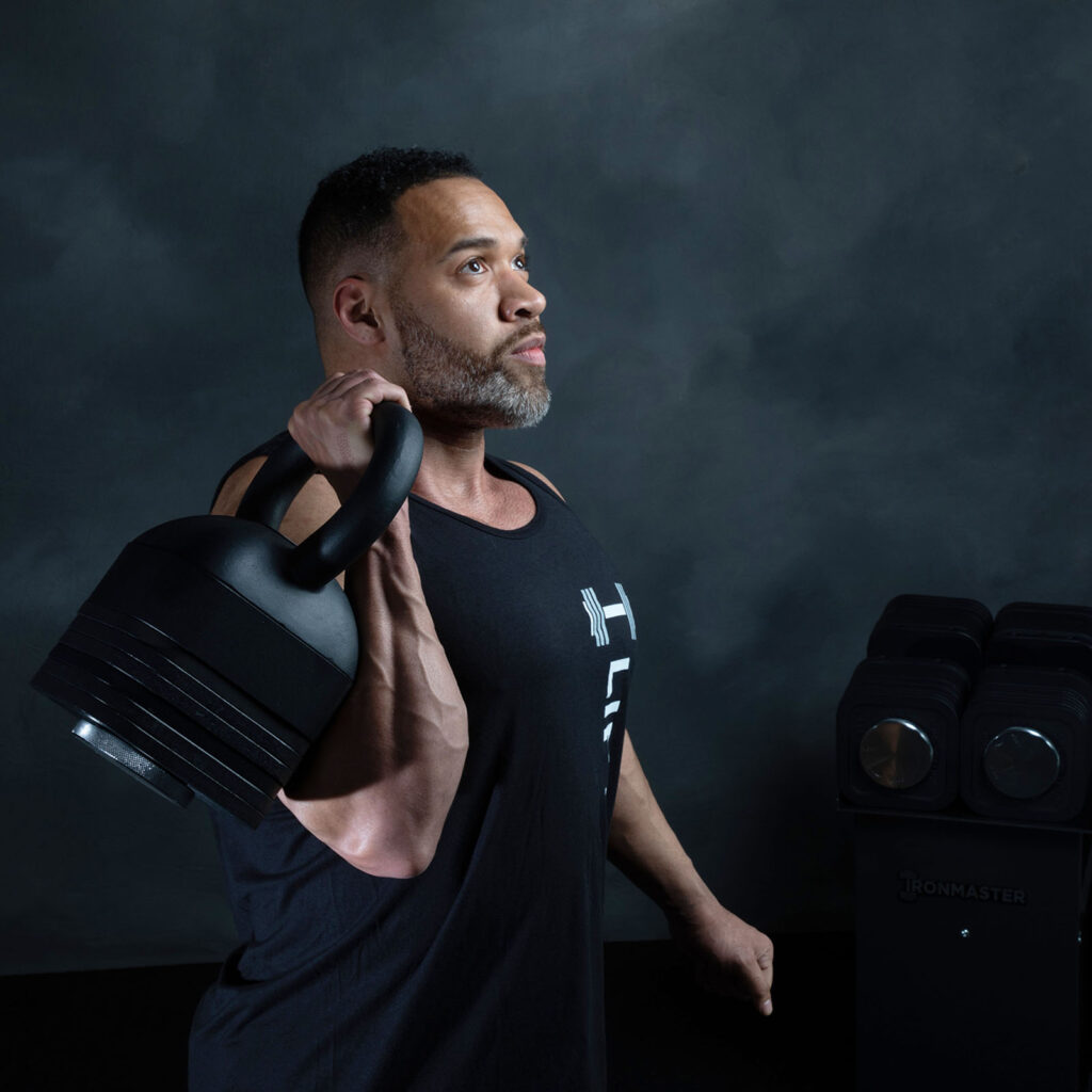 A man in a black tank top holds an Ironmaster Adjustable Kettlebell Handle over his shoulder, looking focused. Behind him, a stack of weights is set against a dark, textured wall.