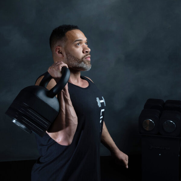 A man in a black tank top holds an Ironmaster Adjustable Kettlebell Handle over his shoulder, looking focused. Behind him, a stack of weights is set against a dark, textured wall.