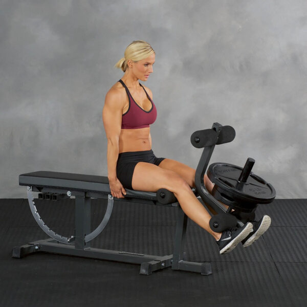 Dressed in workout gear, an individual uses the Ironmaster Leg Extension Attachment on a gym machine. Seated on a bench, they extend their legs against the weights. The setting remains a plain gray background.