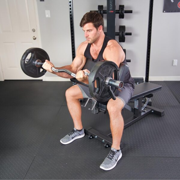 In a gym setting, a man in a black tank top and gray shorts does bicep curls with an EZ bar on the Ironmaster Preacher Pad, surrounded by weights and equipment.