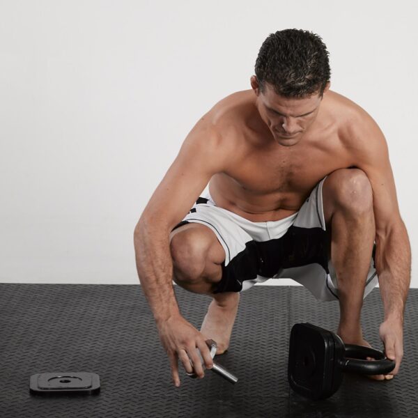 A shirtless man, in athletic shorts, squats on a black mat adjusting a Standard Kettlebell Locking Screw while a weight plate lies nearby.