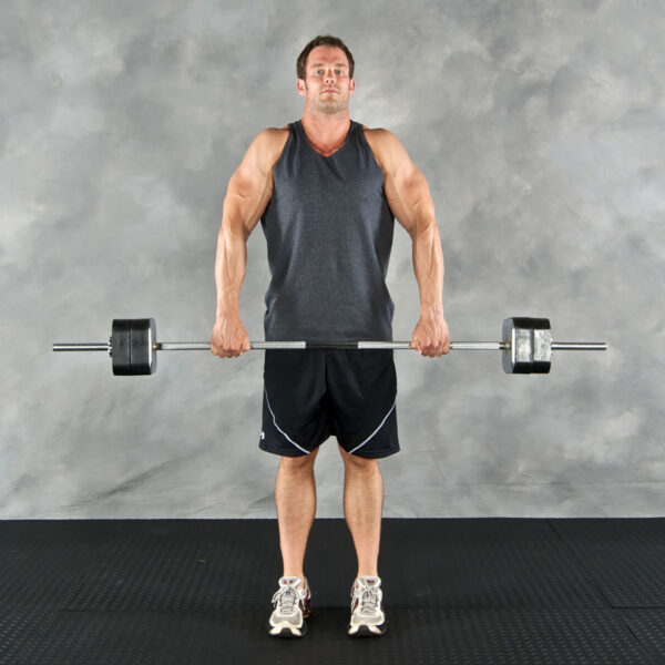 A man in a dark sleeveless shirt and black shorts uses an Ironmaster Straight Barbell to perform an upright row on the gyms black floor against a textured gray background.