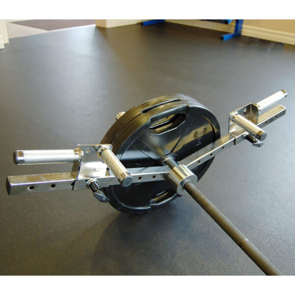 A gym setup features a loaded trap barbell with black weight plates on a rubber gym floor. The Ironmaster Ultimate Row Handle, in chrome, is ready for deadlifts or shrugs. Gym equipment and part of a doorway are visible in the background.