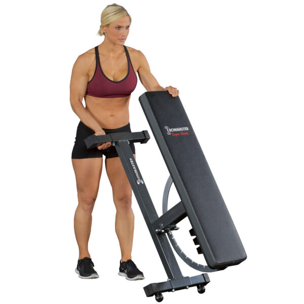 A person in maroon sports bra and black shorts adjusts the Ironmaster Wheelkit for Super Bench, a folding incline workout bench in black equipped with wheels.