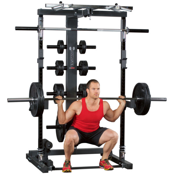 Wearing a red tank top and black shorts, a person is squatting with a barbell on the Ironmaster IM2000 Spotter Arms, which have additional weight plates. The person is focused and using proper form throughout the exercise.