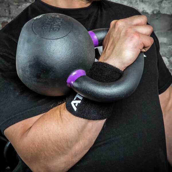 A person clutches a large black kettlebell accented with purple, wearing a Kettlebell Wrist Guard. The backdrop is a textured gray wall.