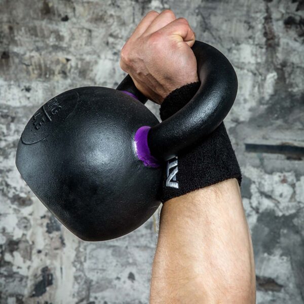 Close-up of a person lifting a black kettlebell, wearing a Kettlebell Wrist Guard. The textured concrete wall serves as the backdrop.