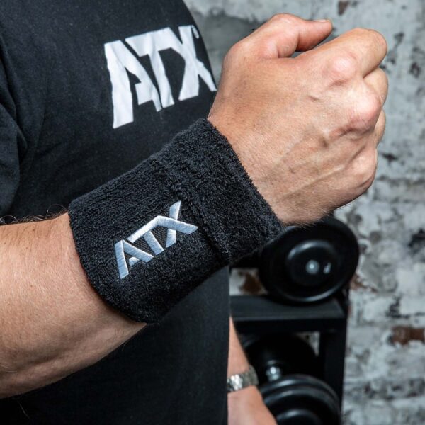 A person wearing a Kettlebell Wrist Guard with ATX embroidered on it raises their fist, paired with a black t-shirt featuring the same logo. Blurred in the background are weights and a brick wall.
