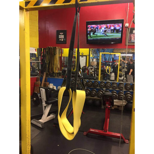 A gym scene features Spud Incs The Big Ray Strap in yellow hanging in the foreground. In the background, weightlifting equipment, dumbbell racks, and a TV showing a sports game are visible. The gym has bright yellow walls with a red accent wall.