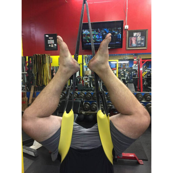 In a gym with red and yellow walls, weights in the background, and a TV on the wall, a person is upside down on an inversion table using Spud Incs The Big Ray Strap to secure their feet.