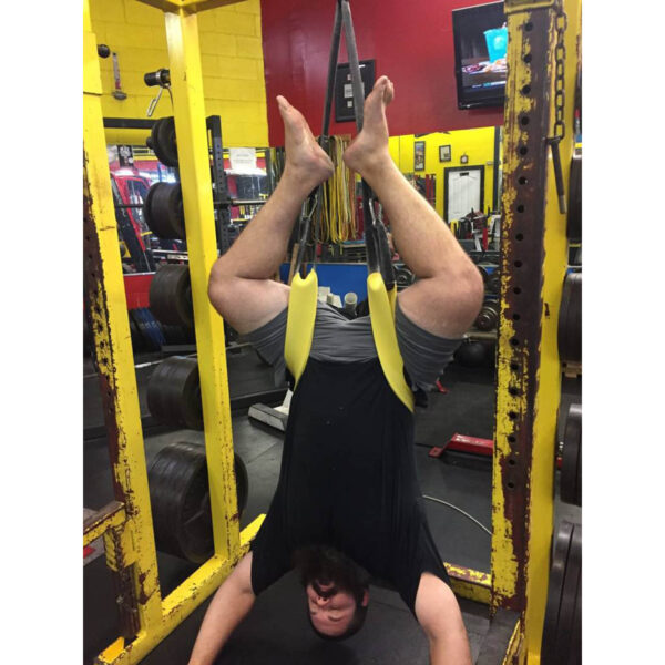 A person hangs upside down in a gym using Spud Inc. The Big Ray Strap wrapped around their legs, inside a power rack with various gym equipment visible in the background.