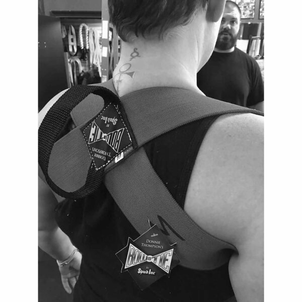 Someone is wearing a Spud Inc Bowtie Casual shoulder support strap, while another person stands in the background. Its a black and white image featuring gym equipment and attire.