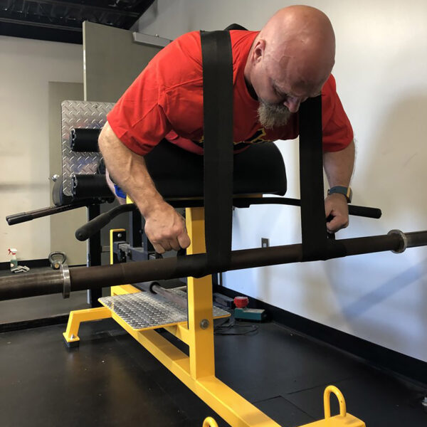 A bald man with a goatee is exercising on a yellow and black hyperextension machine at the gym, wearing a red shirt. He holds Spud Inc Dr Squat Straps, maintaining a focused expression.