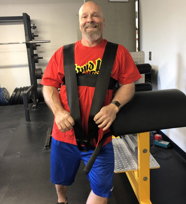In a gym, a bald man with a beard smiles while wearing a red shirt and blue shorts. Hes using the Spud Inc Dr Squat Strap, which is black and attached to gym equipment. Weights are visible in the background.