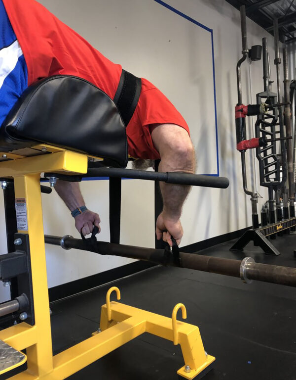A person in a red shirt is using a Spud Inc Dr Squat Strap in the gym. They lie face down, holding a barbell with their right hand, bending at the waist. Various weights and equipment are visible in the background.