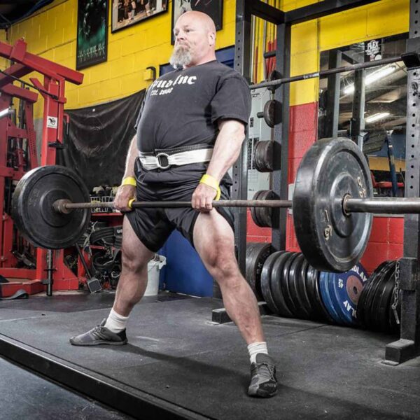 In a vibrant gym with colorful walls, someone is deadlifting in a black t-shirt, shorts, and weightlifting belt while wearing Spud Elbow Sleeves. Weight plates are neatly stacked behind them.