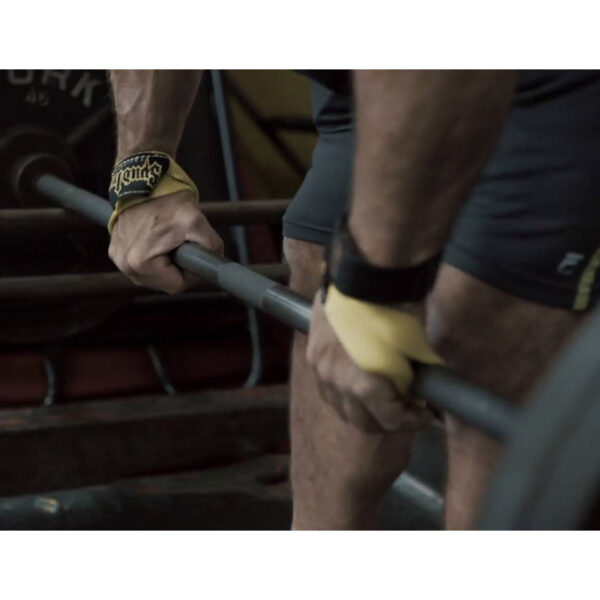 A person grips a barbell using the Spud Inc Fat Triceps Strap, readying for a deadlift in the gym. Their hands and legs are in focus, with gym equipment visible in the background.