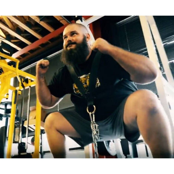 A bearded man is using a Spud Inc. Front Squat Harness for squats in the gym, with equipment and ceiling beams visible in the background.