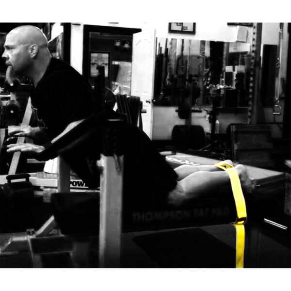 A person exercises on a hyperextension bench in a gym, secured with the Spud Inc Glute Ham Strap around their legs. The gym features various weights and mirrors in the background.