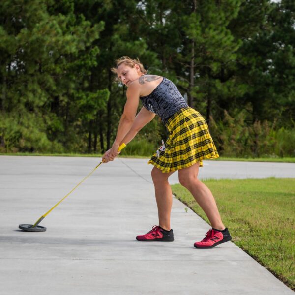 A person in a yellow plaid skirt and black top is holding the Spud Inc Econo Hammer by its yellow rope on a concrete path, with trees and grass in the background.