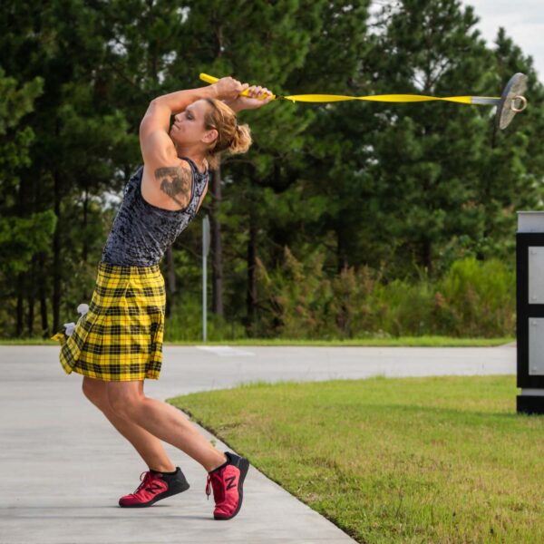 A person in a gray tank top and yellow plaid kilt prepares to throw the Spud Inc Econo Hammer overhead on an outdoor concrete path, with trees in the background, while wearing red athletic shoes.