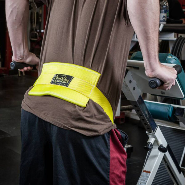 A person in a brown shirt with a yellow Spud Chainless Dip Belt exercises on a dip machine at the gym. Gym equipment and a water bottle are in the background.