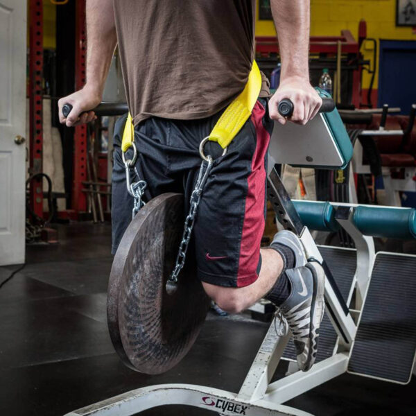 A person is using the Spud Chainless Dip Belt to perform a dip exercise on a machine, securing a large weight plate between their legs. The gyms background equipment adds to the focused workout atmosphere.