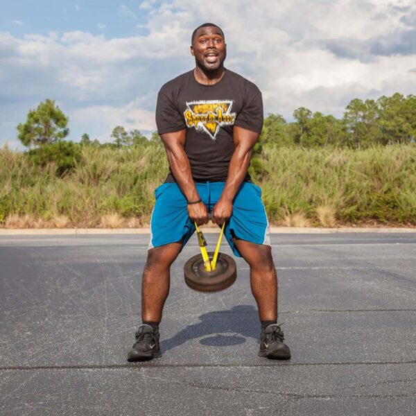 A man using a Spud KettleStrap Kettlebell Strap to lift a weighted sled outdoors. Hes on a paved area, wearing a black T-shirt, blue shorts, and black sneakers, with greenery and a partly cloudy sky in the background.