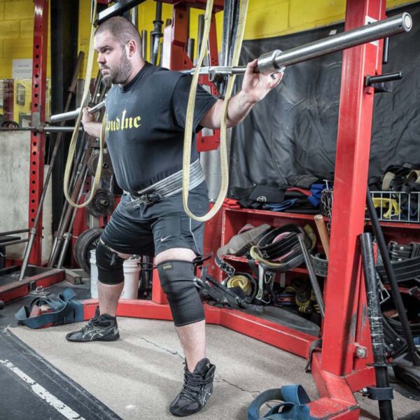 A person in a black T-shirt and Spud Knee Sleeves 1 Ply is doing a squat with a barbell at the gym. Red squat racks, lifting equipment, and weights are visible around them.
