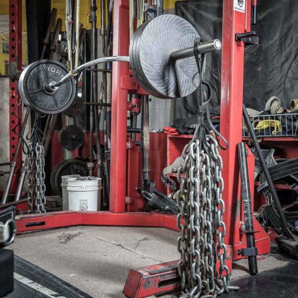A weightlifting area features a red squat rack with a barbell loaded with weights and chains on each end. Various gym equipment, including Spud Looper Straps, fills the area on the concrete floor.