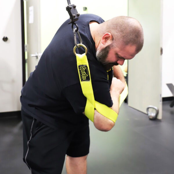 A bearded man with a shaved head uses the Spud Standing Oblique Strap in a gym. Gripping the yellow strap firmly with both hands, he bends forward while attached to a harness and pulley system. The room features white walls and a black floor.